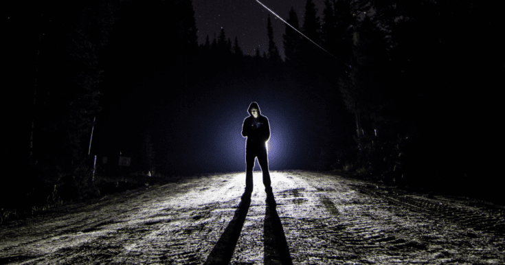 Dude in black pants and black hoodie standing in front of headlights in what appears to be the middle of nowhere.