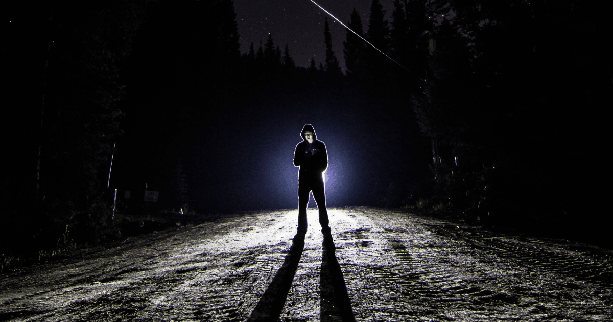 Dude in black pants and black hoodie standing in front of headlights in what appears to be the middle of nowhere.
